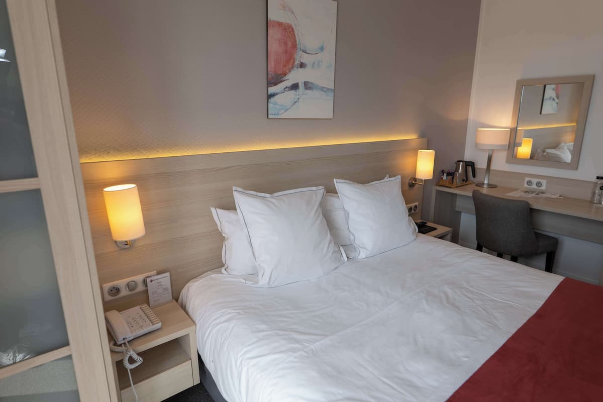 Habitación de hotel con una cama bien hecha, lámparas de noche, un escritorio con una silla, un teléfono y un cuadro en la pared.