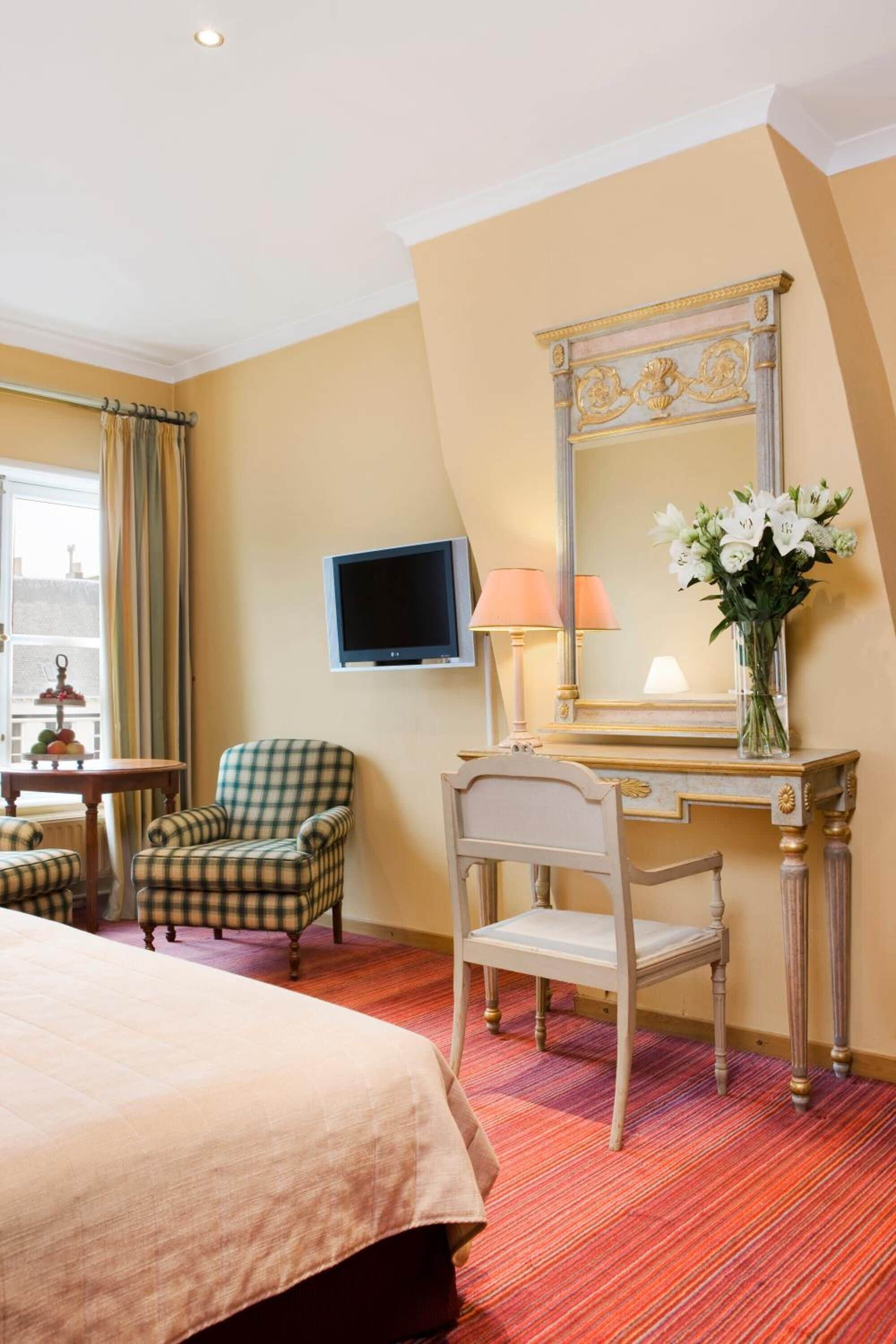 A hotel room with a bed, two chairs, a desk with a mirror, a television, and a vase with flowers.