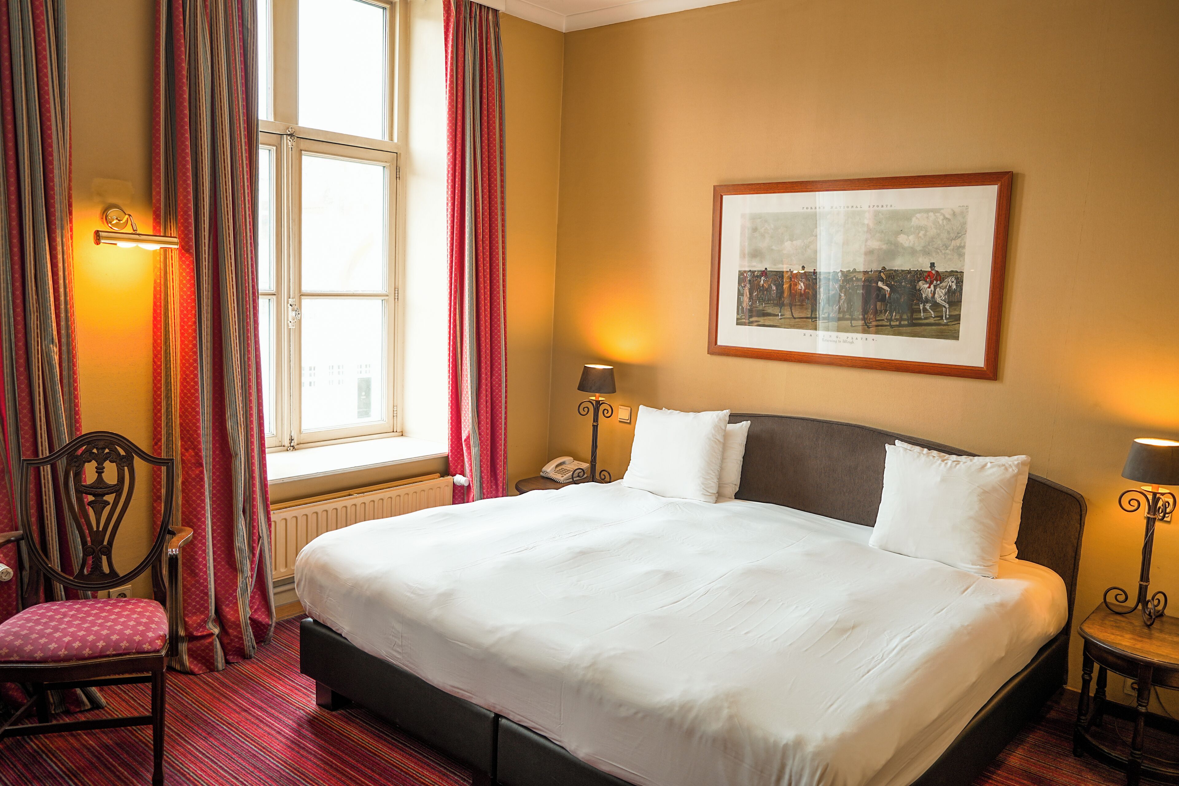 A hotel room with a large bed, a chair, a bedside table with a lamp, a framed picture on the wall, and a window with curtains.