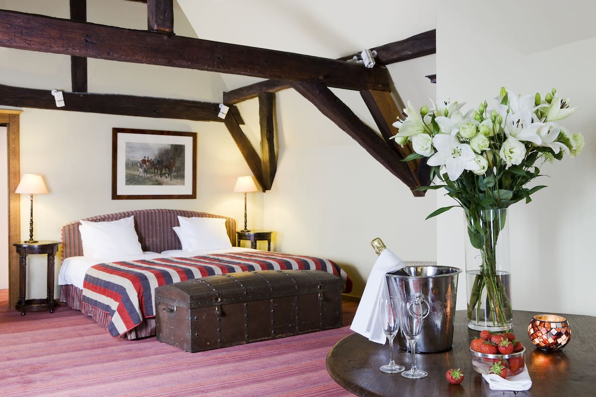 Un dormitorio con una cama, un banco de madera, un jarrón con flores, un balde y una vela sobre una mesa.