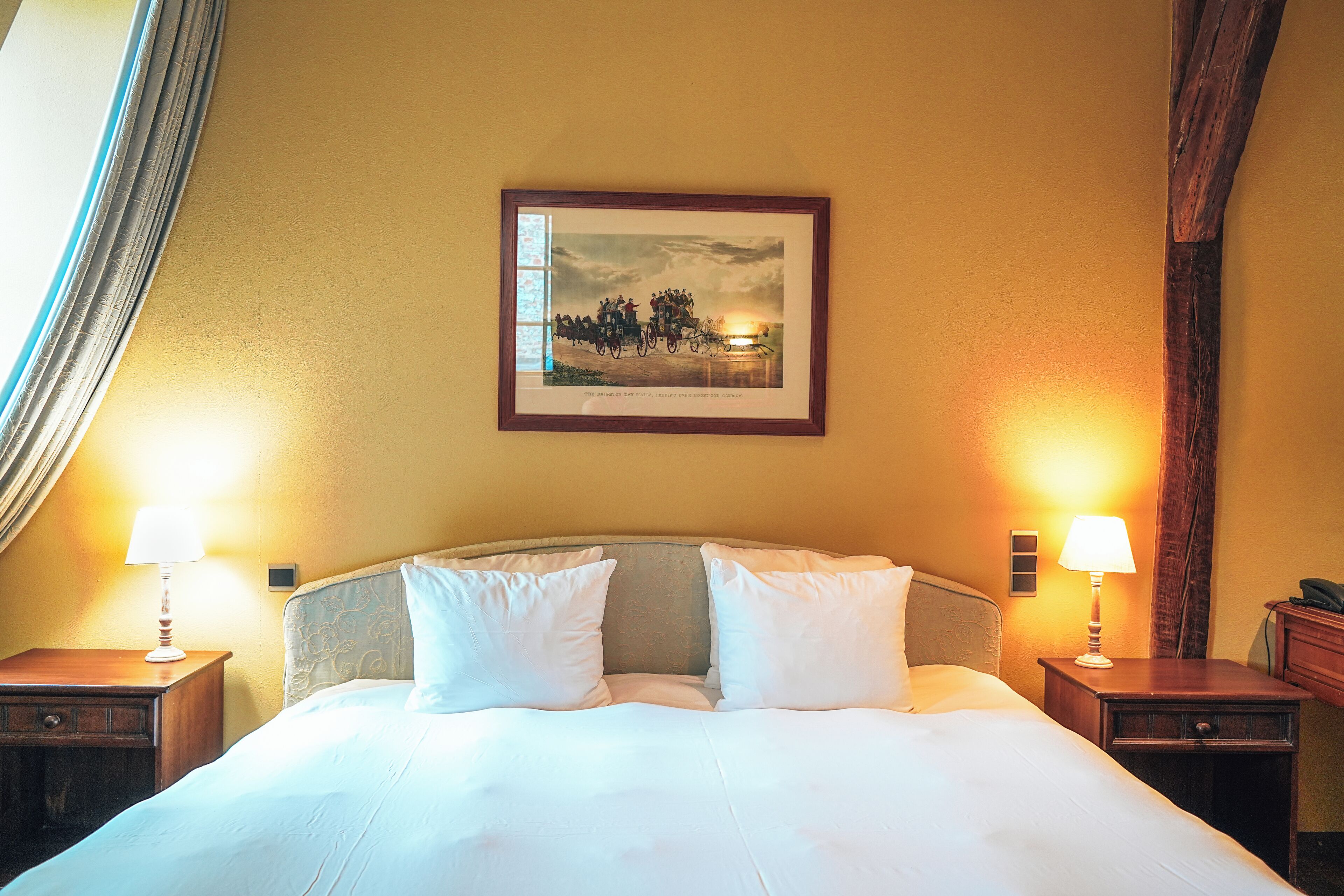 Une chambre d’hôtel avec un grand lit, deux tables de chevet avec des lampes, un tableau encadré au mur et une fenêtre avec des rideaux.