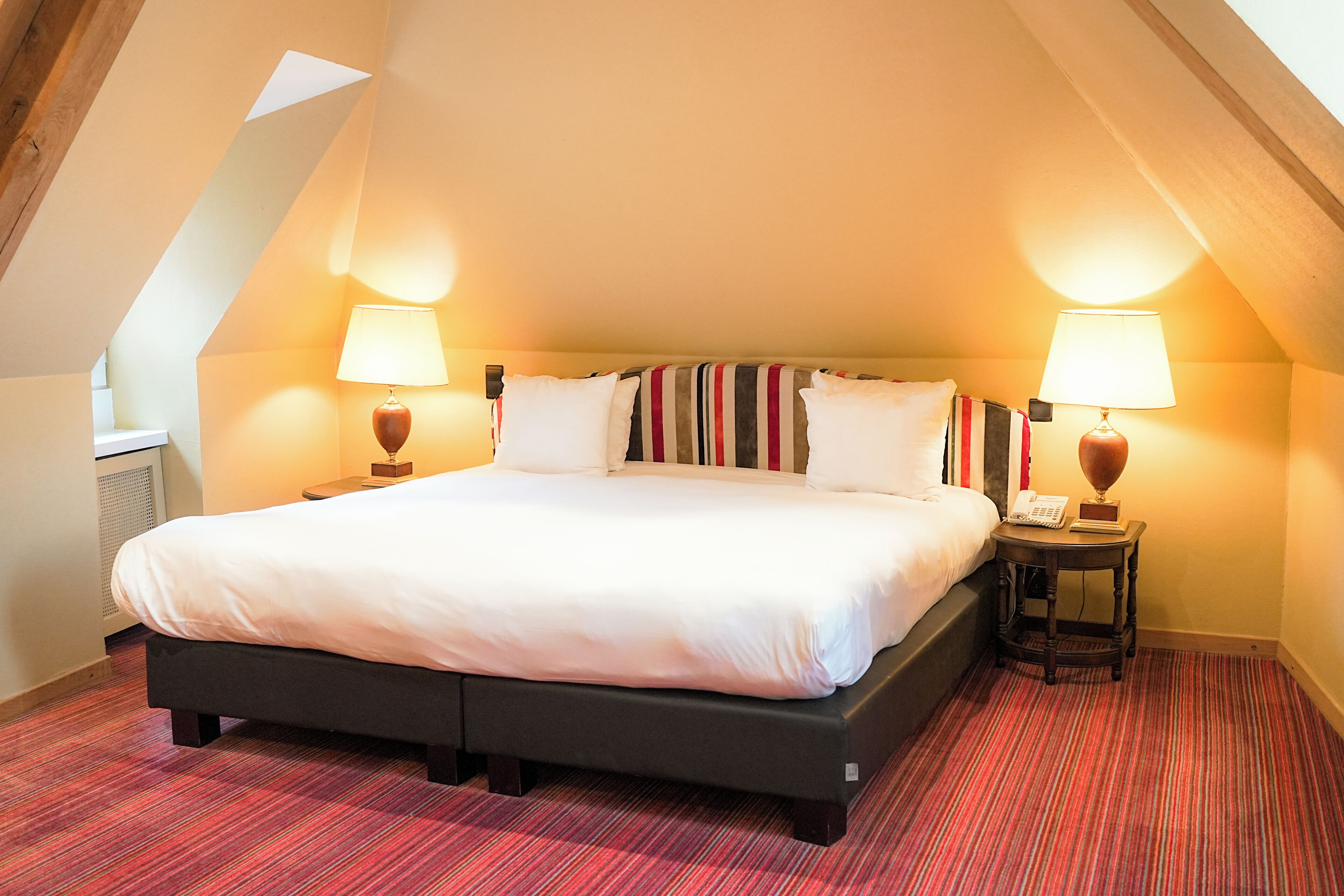 A large bed with white linens in a room with red carpet, two bedside lamps, and a telephone on a small table beside the bed.