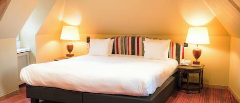 A large bed with white linens in a room with red carpet, two bedside lamps, and a telephone on a small table beside the bed.