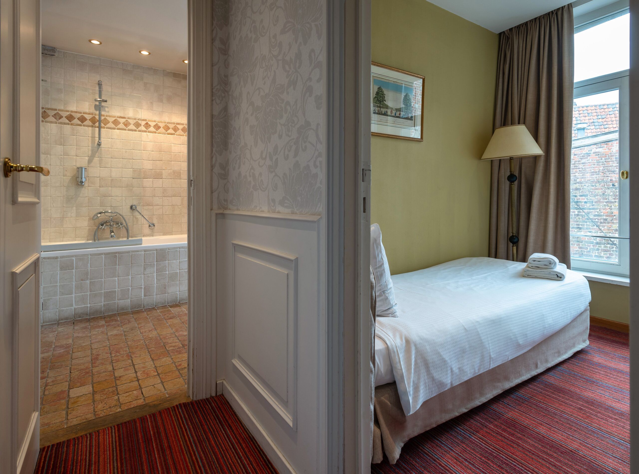 Hotelkamer met een badkamer met bad en douche, een bed met net opgemaakt bed, een raam met gordijnen en een ingelijst schilderij aan de muur.