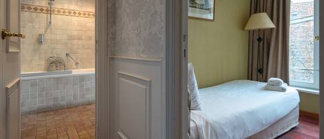 Habitación de hotel con baño que incluye bañera y ducha, una cama con ropa de cama bien tendida, una ventana con cortinas y un cuadro en la pared.