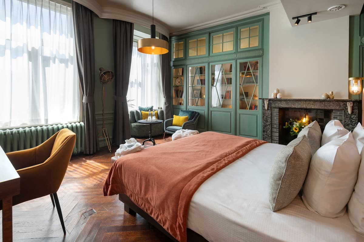 Un dormitorio bien iluminado con una cama grande, un escritorio y una silla. Hay una chimenea y una estantería en la habitación.
