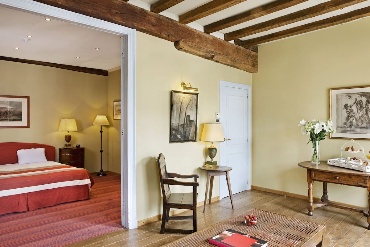 Una habitación con una cama, una silla de madera, una mesita con un jarrón de flores y un cuadro en la pared.
