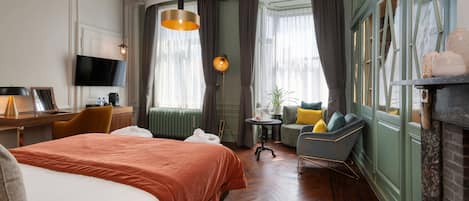 Une chambre d’hôtel avec un grand lit, un bureau avec un ordinateur, une télévision, une cheminée et une vue sur la ville par les fenêtres.