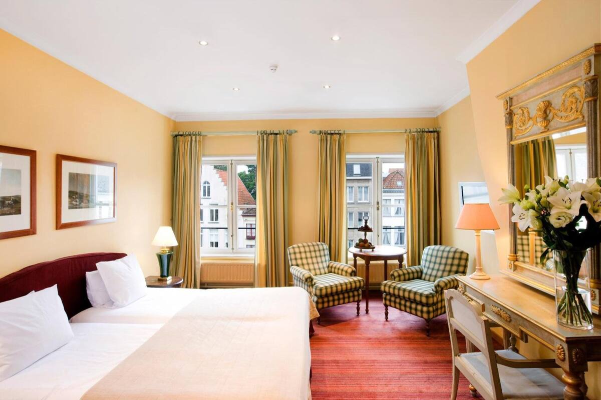 Habitación de hotel con una cama, dos sillas, un escritorio, una lámpara, un florero con flores y un espejo grande.