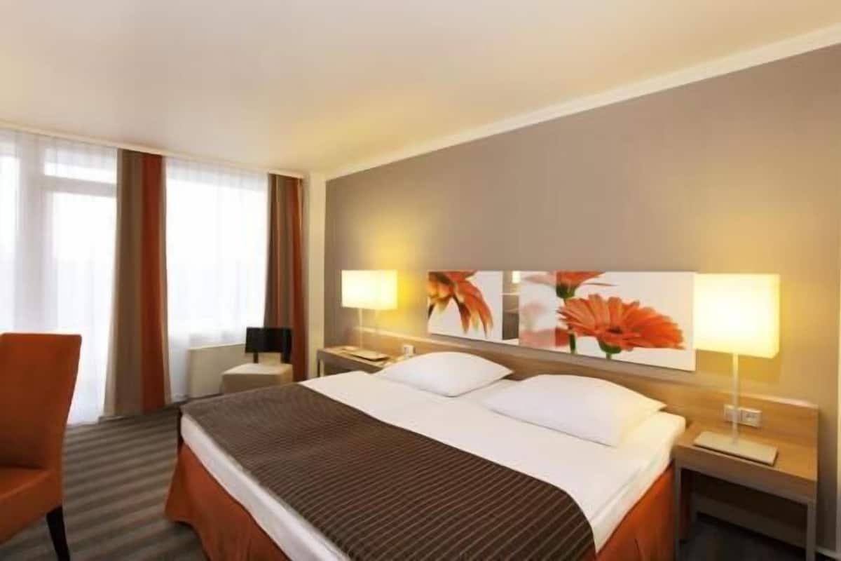 Ein Hotelzimmer mit einem Bett, zwei Nachttischen, einem Sessel, einem Fenster mit Vorhängen und einer Wand mit zwei Blumenbildern.