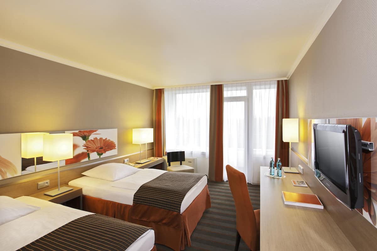 Ein Hotelzimmer mit zwei Betten, einem Schreibtisch mit Fernseher, einem Spiegel und einem Fenster mit Vorhängen.