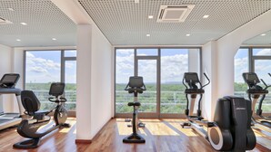 Ein Fitnessraum mit mehreren Ergometer und Laufbändern, großen Fenstern mit Blick auf Grünflächen und einem Holzboden.