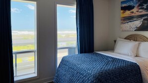 A bedroom with a bed, a painting of a beach scene, and a view of the ocean through large windows. - Mariner Inn and Suites (Kill Devil Hills)