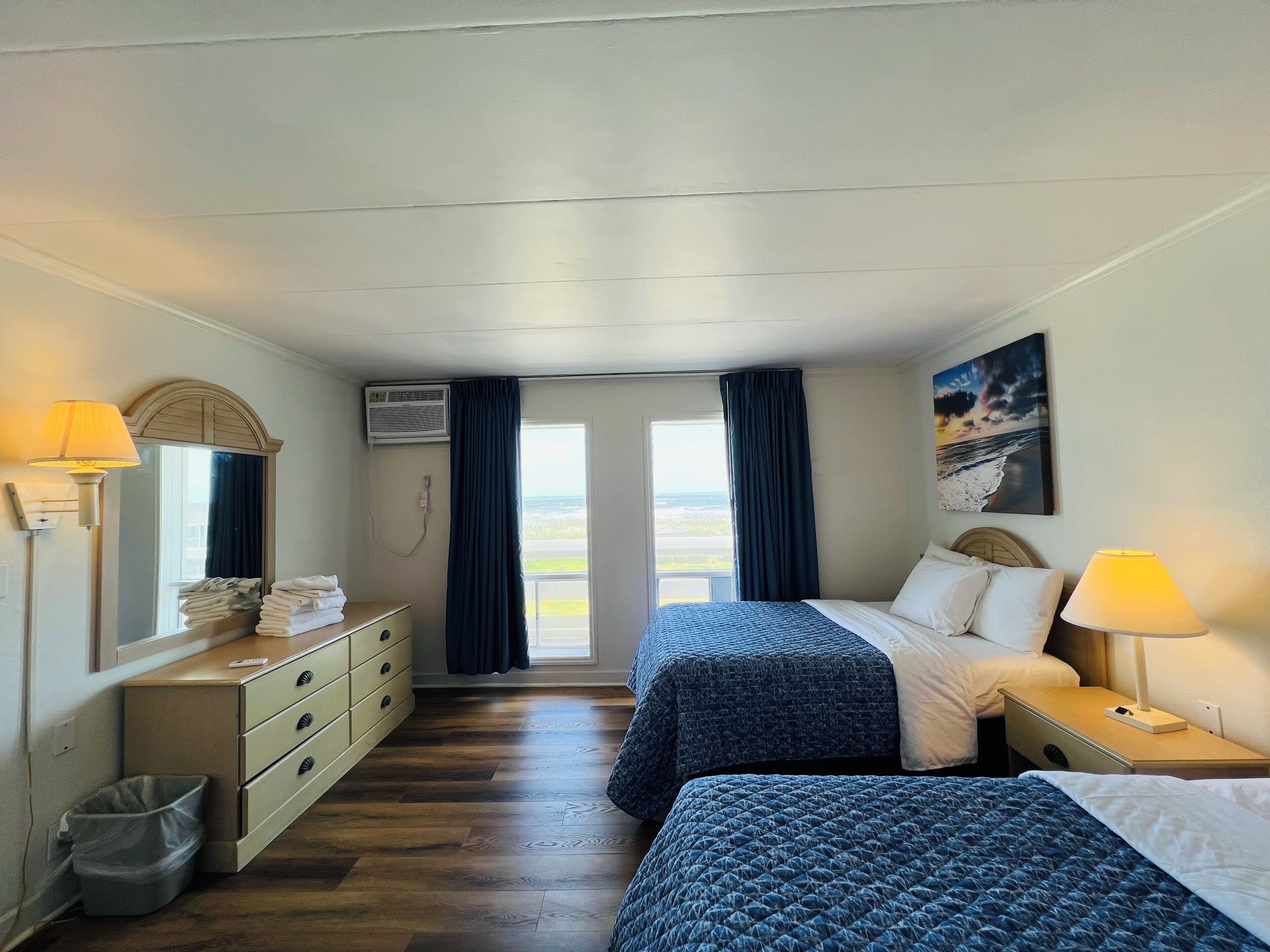 A hotel room with two beds, a dresser, a trash bin, and a view of the ocean.