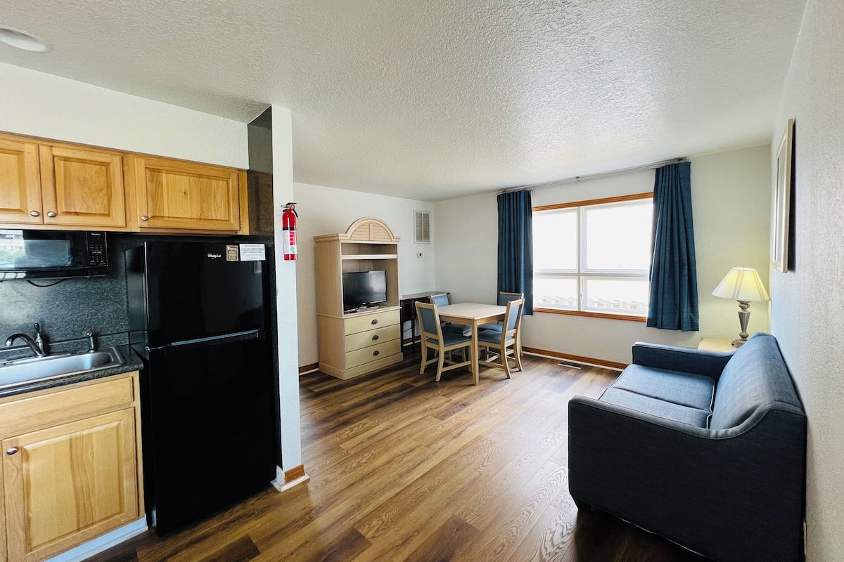 A compact kitchen with wooden cabinets, a black refrigerator, and a microwave. There's a dining area with a table and chairs, and a sofa.
