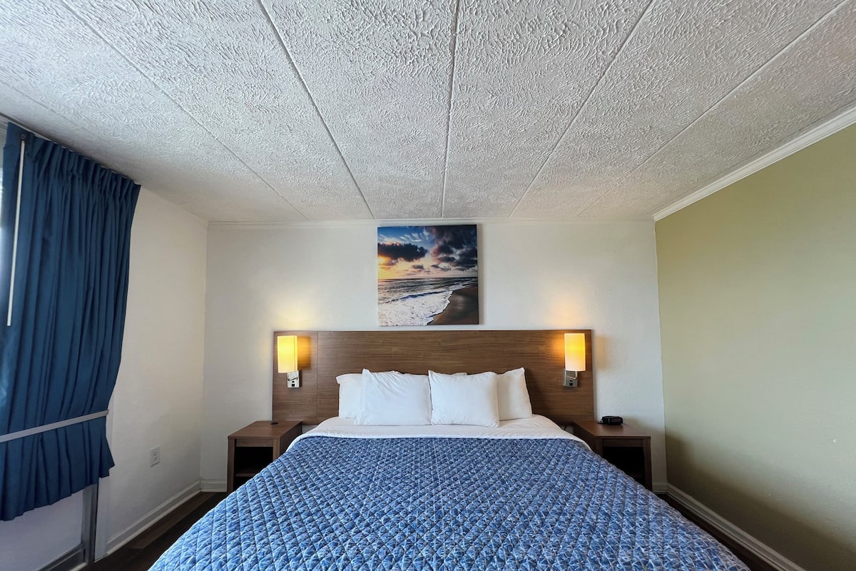 A hotel room with a bed, two bedside tables, a headboard, a painting of a beach scene, and a window with curtains.
