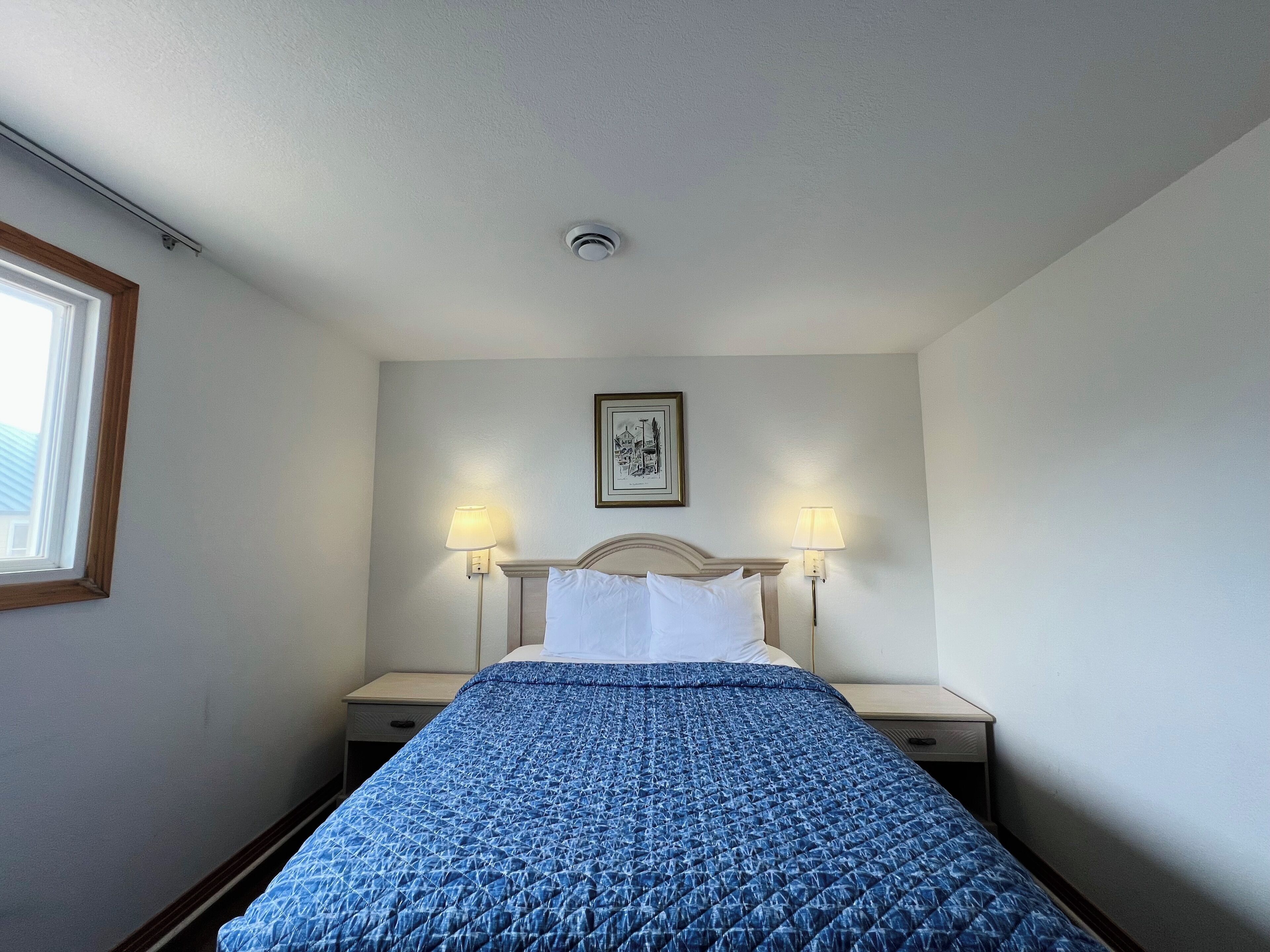 Quarto de hotel com uma cama, duas luminárias de apoio, um quadro na parede e uma janela no lado esquerdo.