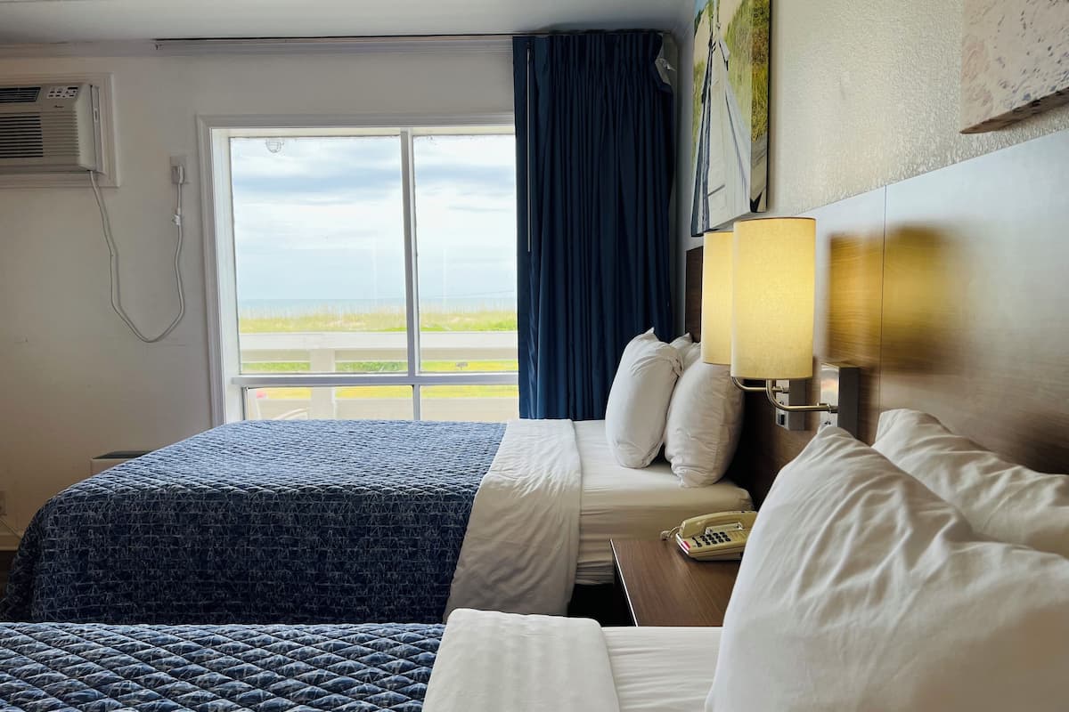 A hotel room with two beds, a window with a view of the beach, an air conditioning unit, and a bedside lamp.