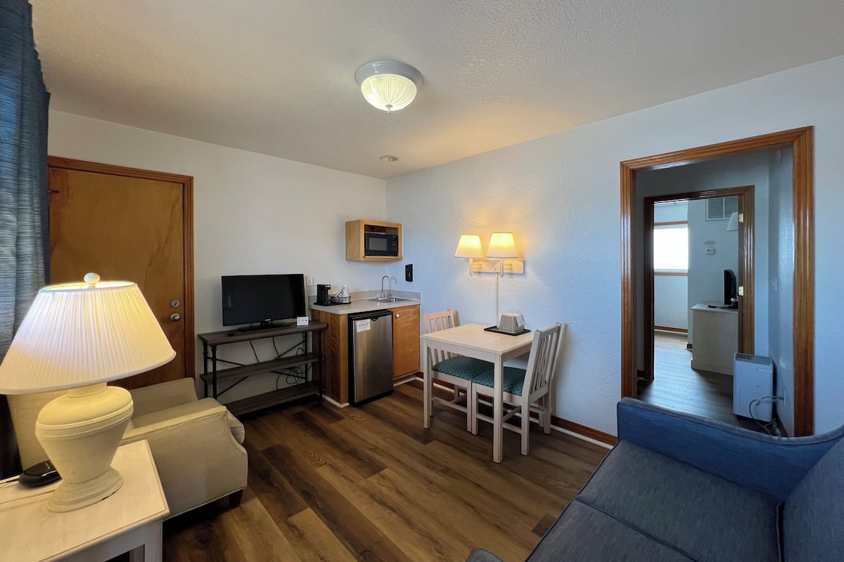 A compact hotel room with a kitchenette, a dining area, a sofa, and a television.