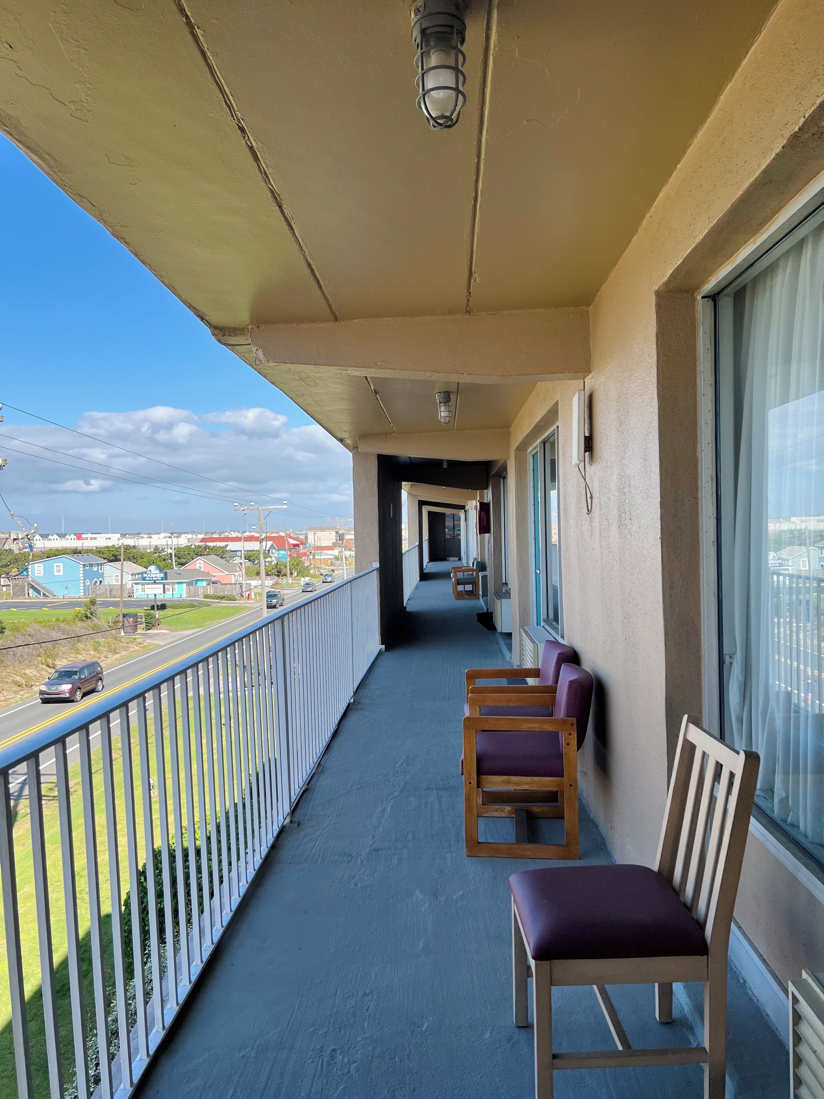 A balcony with a view of a road and buildings, wooden chairs, and a railing.