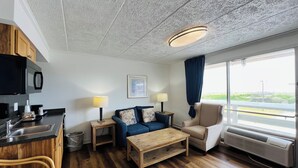 A modern room with a kitchenette, a sofa, a chair, a wooden coffee table, and a large window with a view. - Mariner Inn and Suites (Kill Devil Hills)
