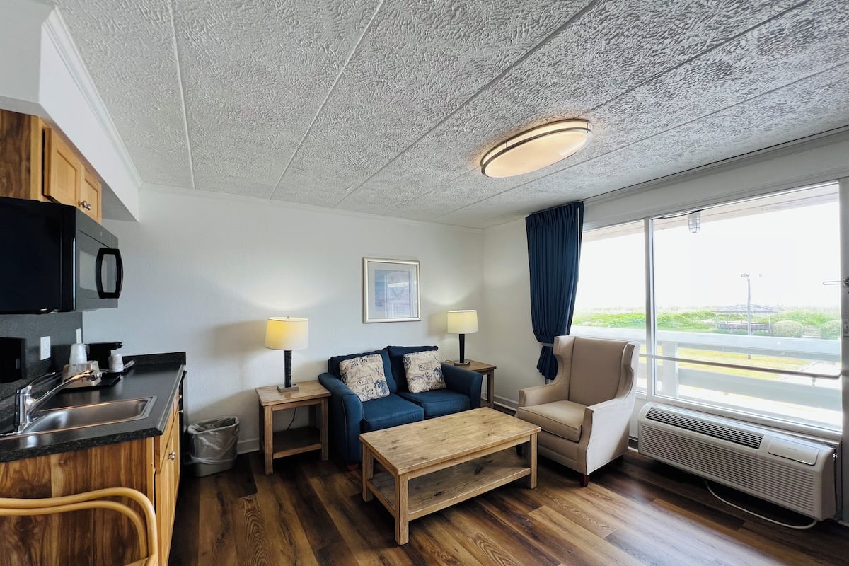 A modern room with a kitchenette, a sofa, a chair, a wooden coffee table, and a large window with a view.