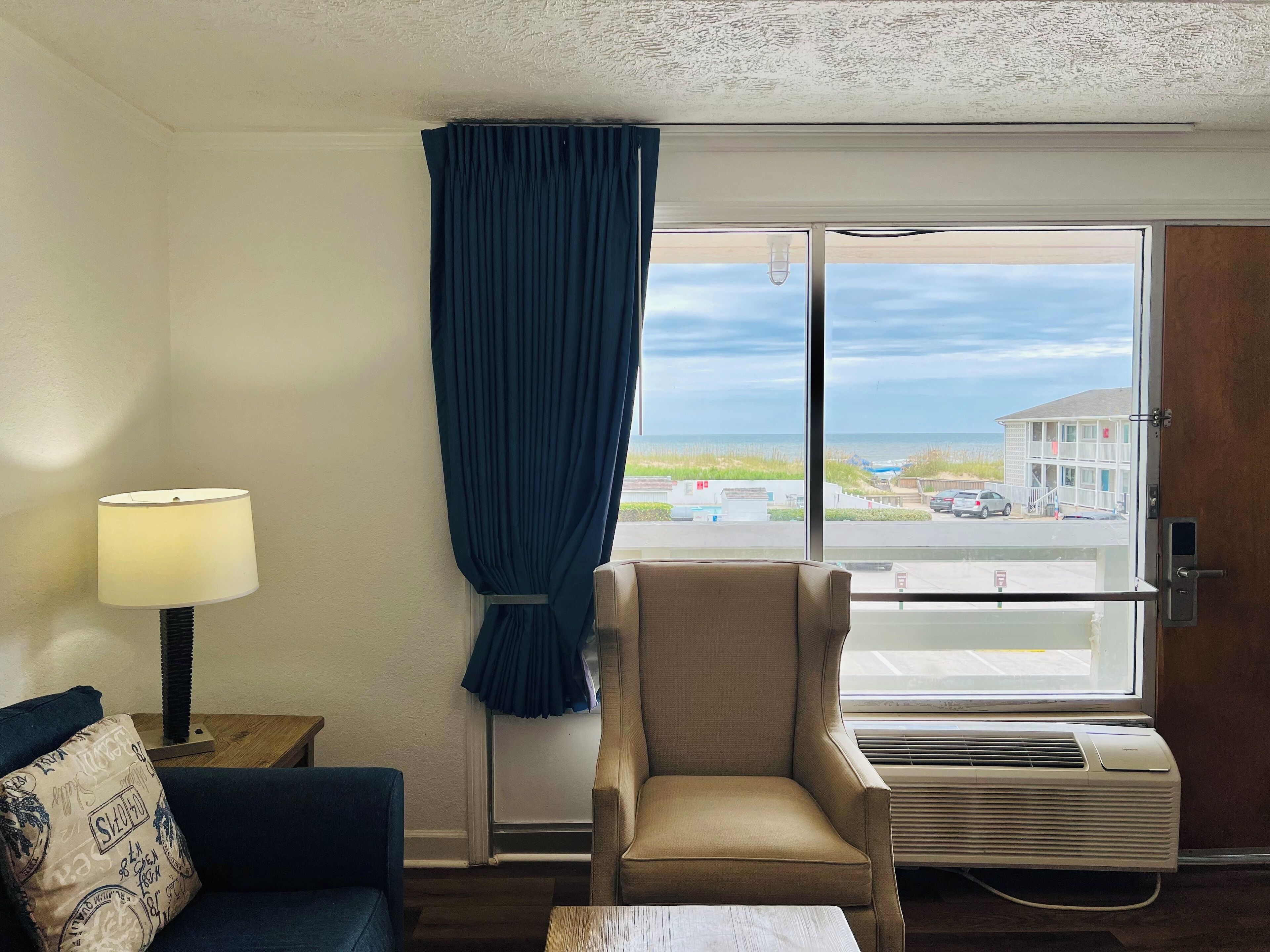 Quarto de hotel com uma janela grande, um sofá, uma cadeira, um abajur e um ar condicionado.