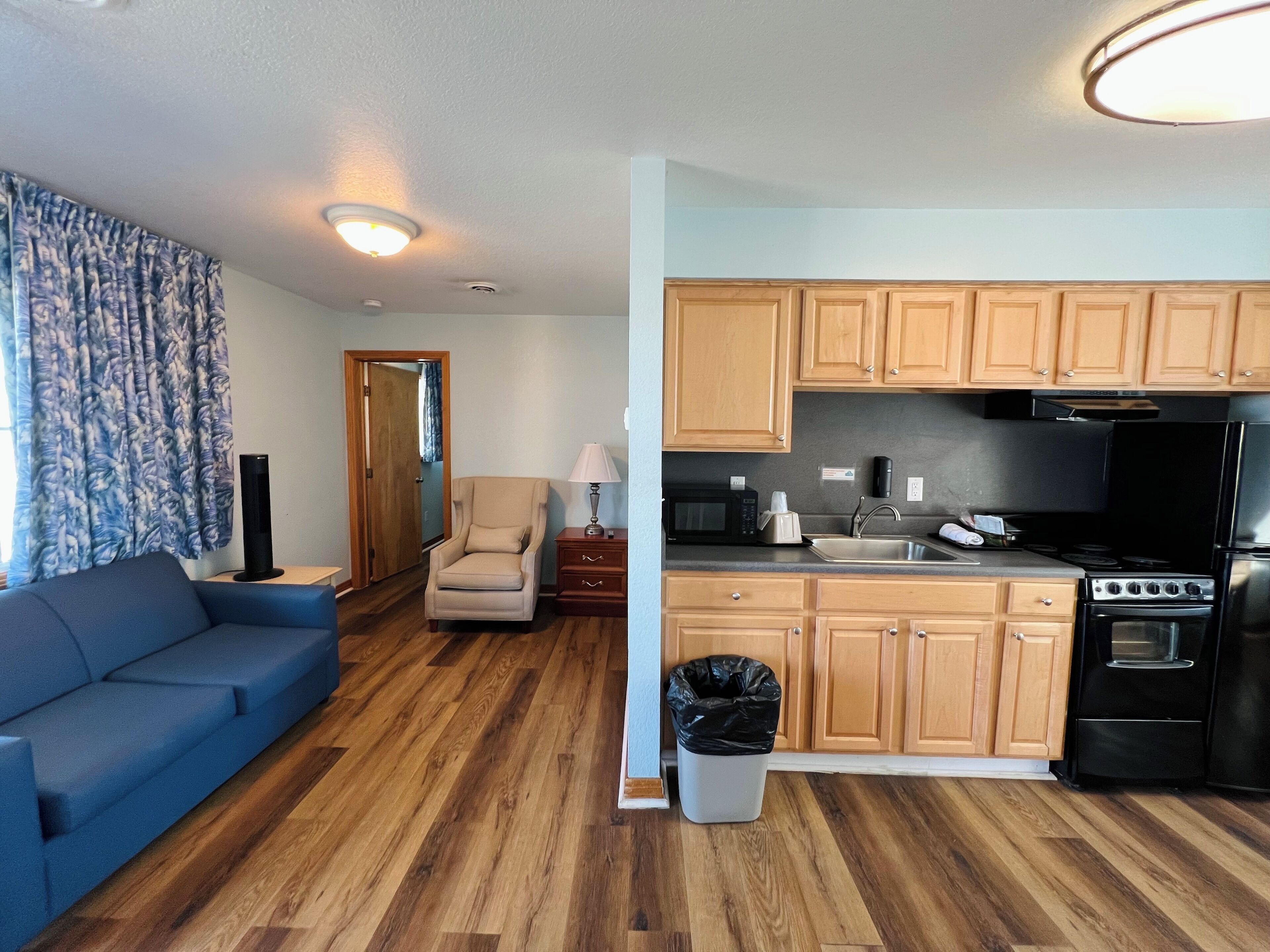 A living room with a blue sofa, a beige armchair, and a small table with a lamp. The kitchen has wooden cabinets, a black refrigerator, and a stove.