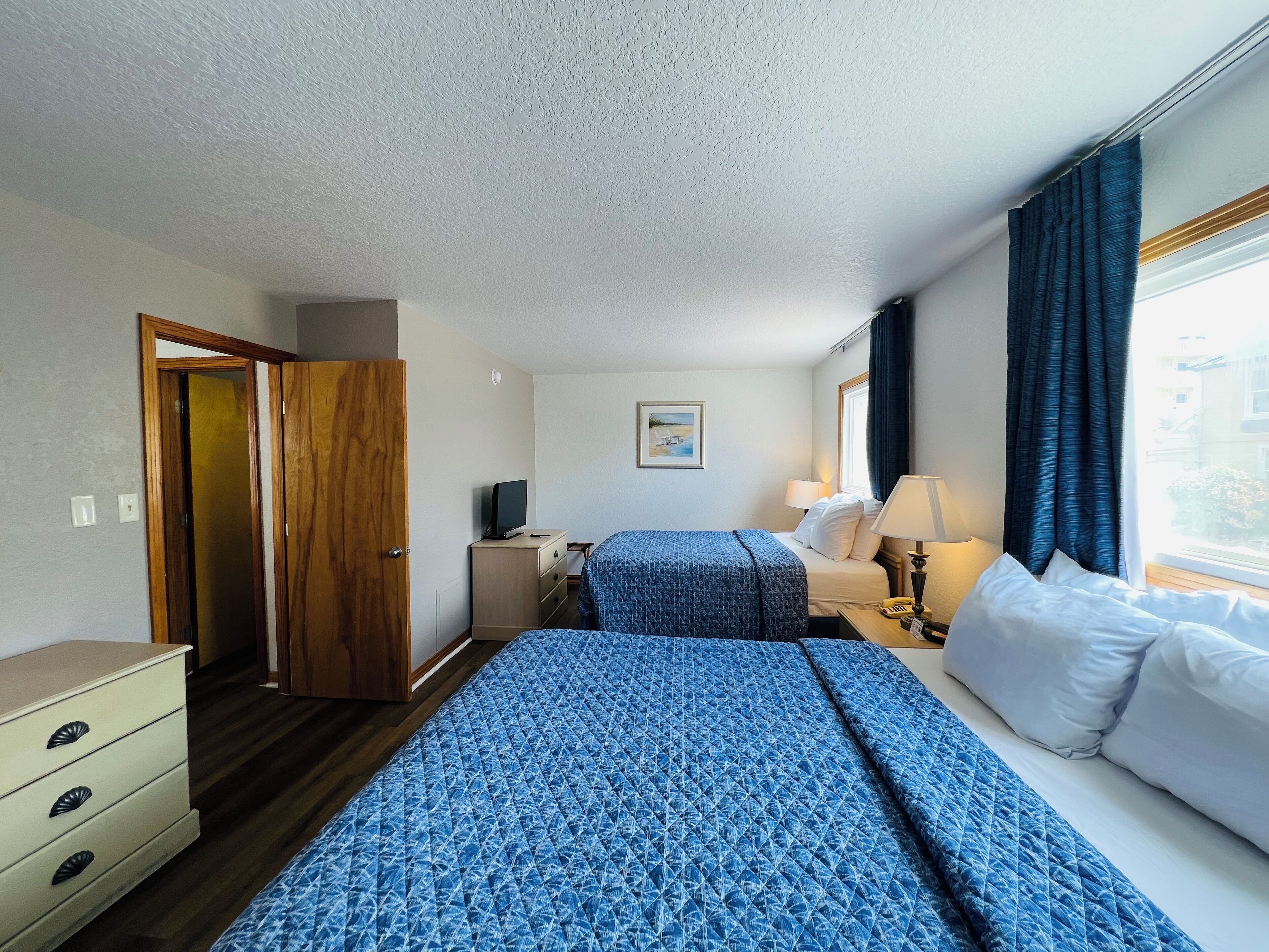 Una habitación de hotel con una cama grande, una cama más pequeña, un escritorio con lámpara, un aparador y una ventana con cortinas.