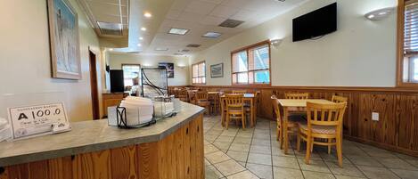 A restaurant interior with a counter, wooden chairs, tiled flooring, and a television on the wall.