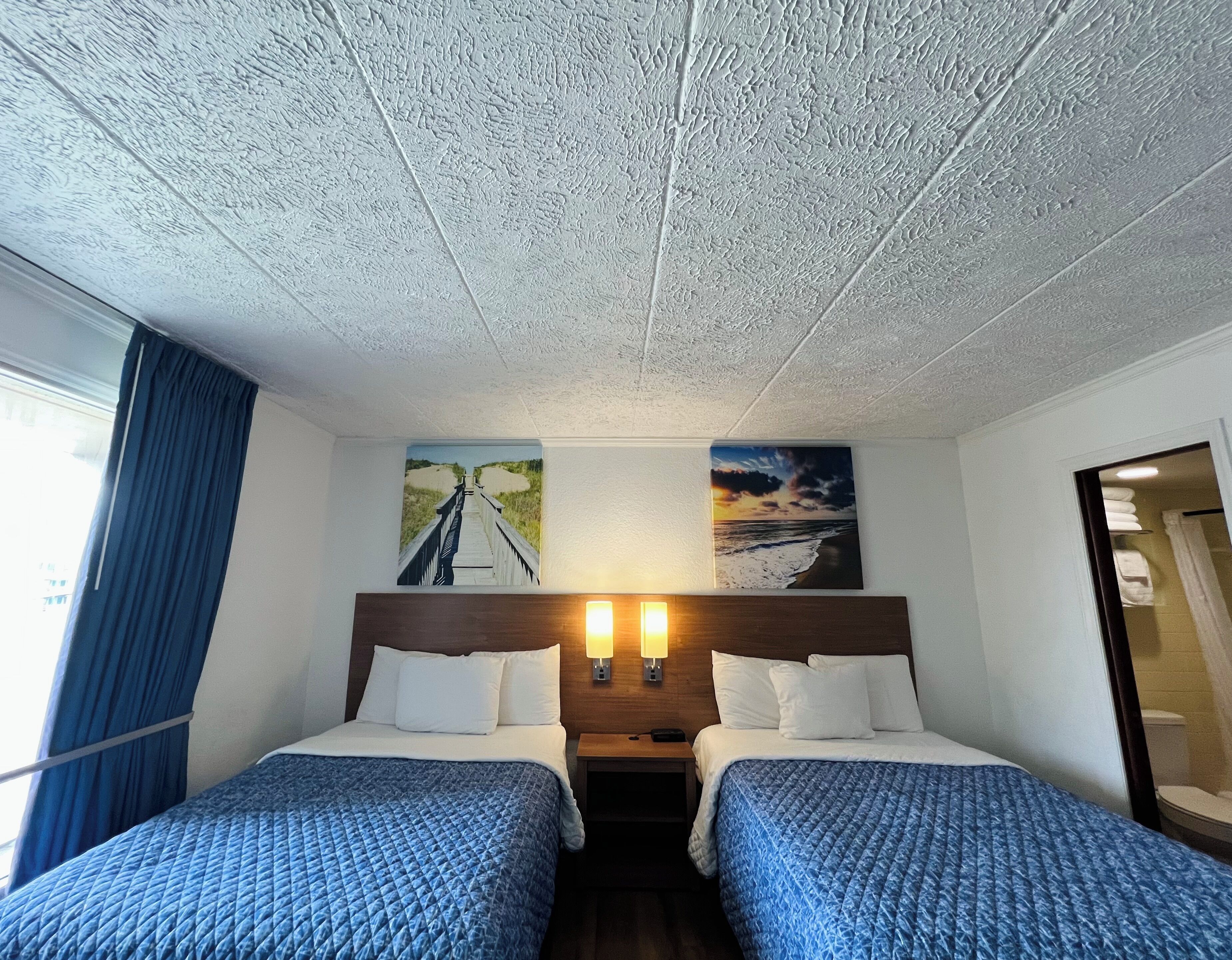 Hotellihuone, jossa on kaksi sänkyä, puinen pääty, yöpöytä ja seinään kiinnitetty lamppu.