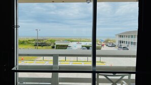 A view from a room with a large window overlooking a beach, a parking area, and a building with a balcony. - Mariner Inn and Suites (Kill Devil Hills)