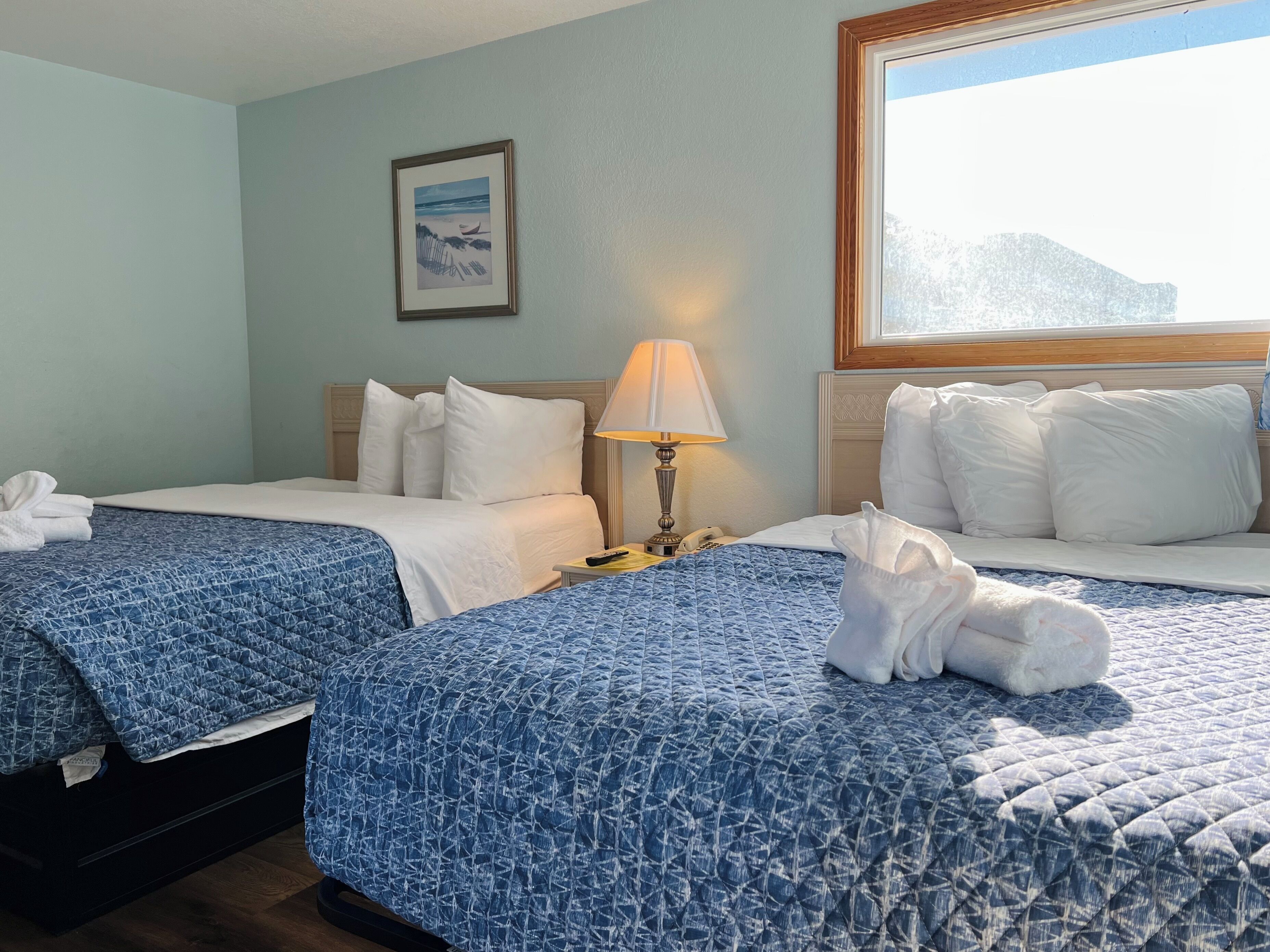 A hotel room with two beds, each with a blue quilted bedspread, white pillows, and a framed picture on the wall.