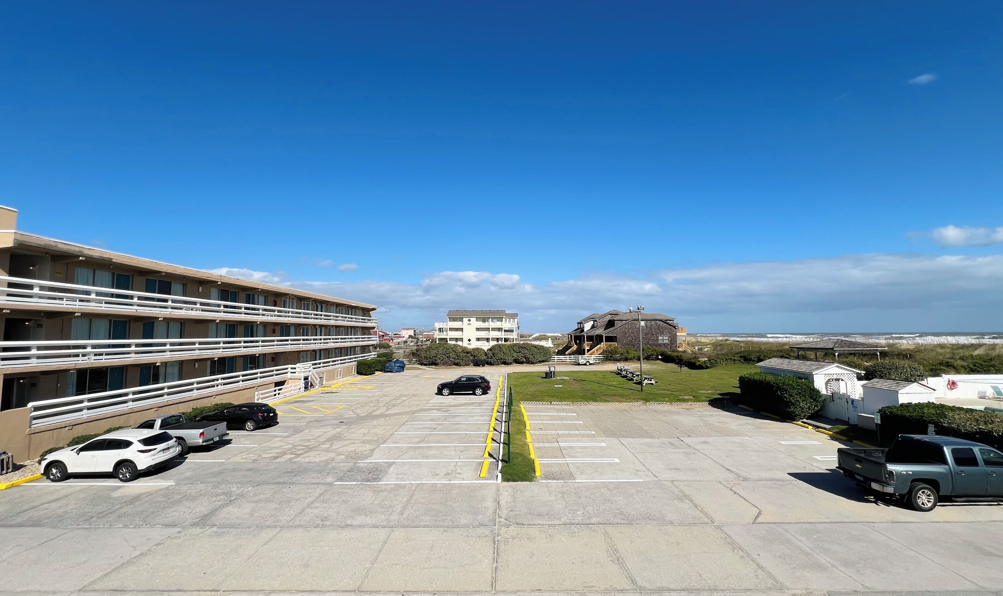 A parking lot with several cars, a building with multiple floors, and a view of the beach in the background.