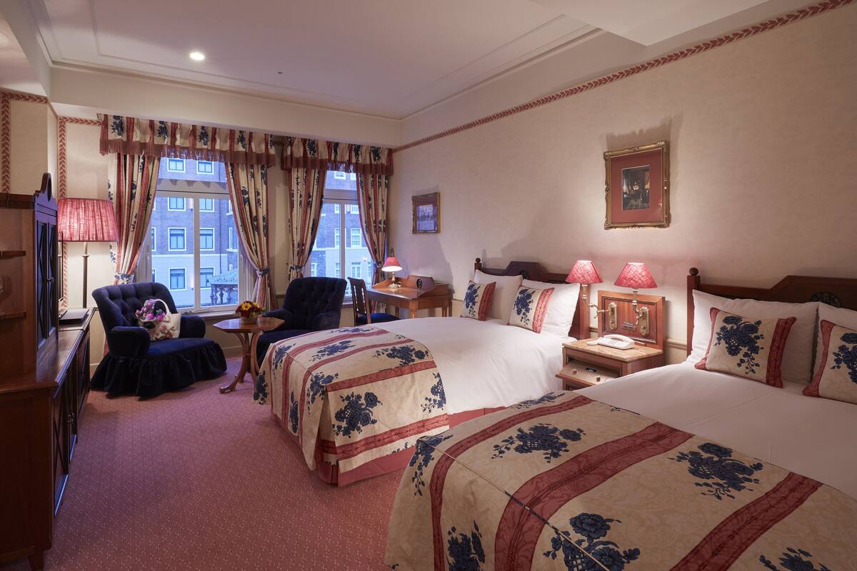 A hotel room with two beds, a desk, a chair, a lamp, and a window with curtains.