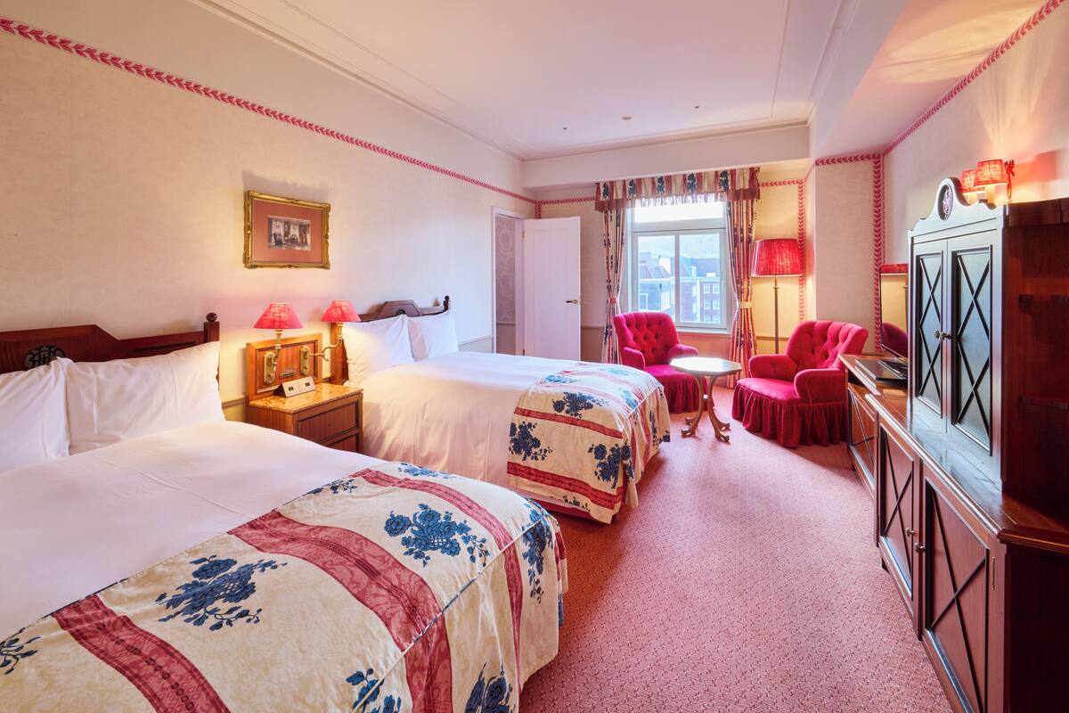 A hotel room with two beds, a red armchair, a small table, and a cabinet.