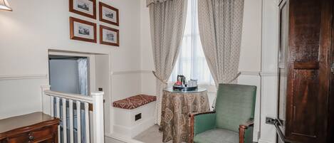 A room with a crib, a green chair, a table with a lamp, and a window with curtains.