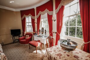 Una habitación con un ventanal grande, un tocador con espejo, un sillón rojo, una silla de madera, una mesa con mantel floral y un bol con hielo y botellas.