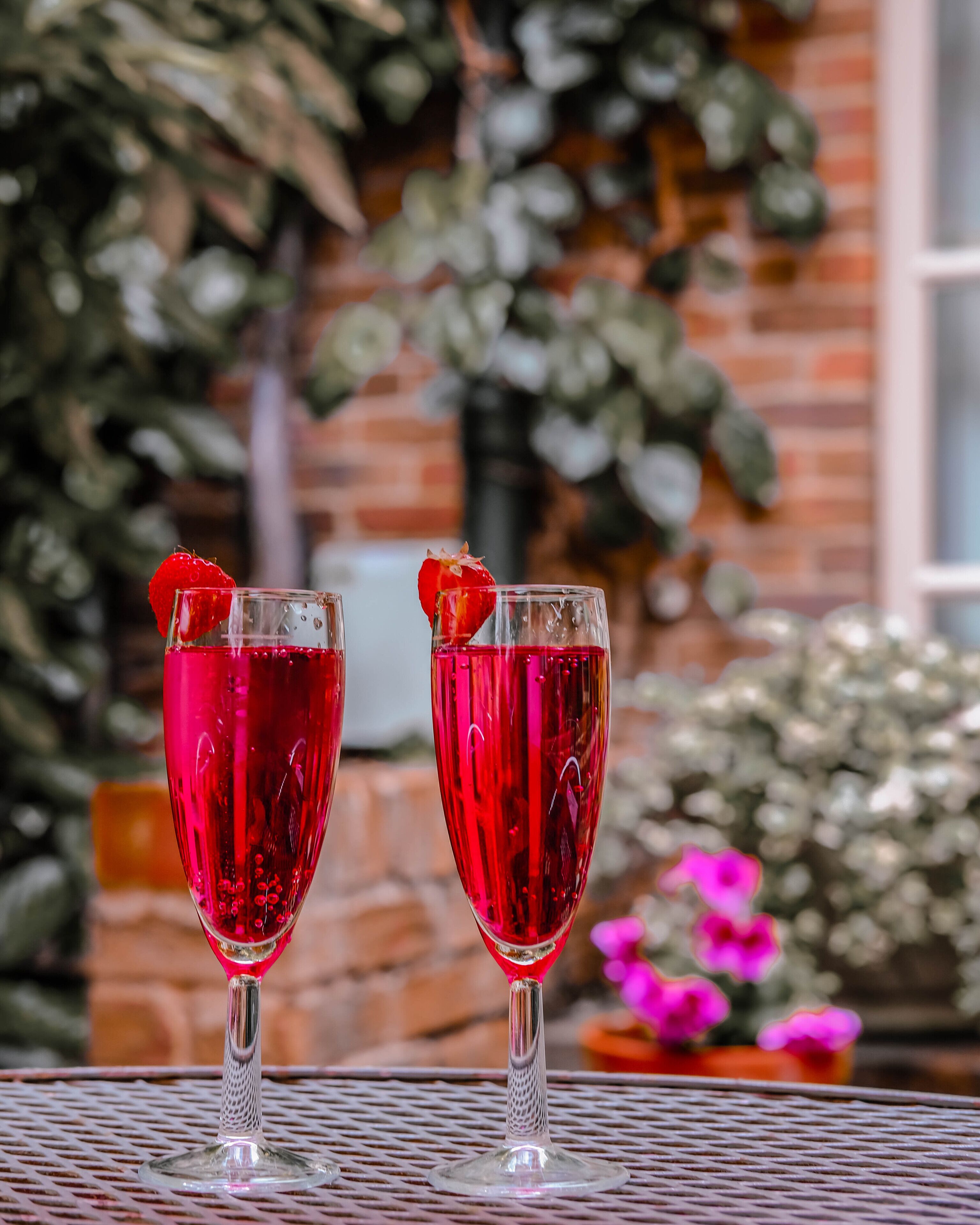 Two glasses of red beverage on a metal table with a brick wall and plants in the background.