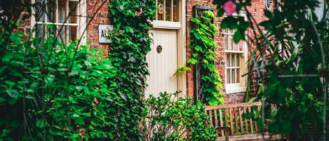 A brick building with a white door, a wooden bench, and greenery around.