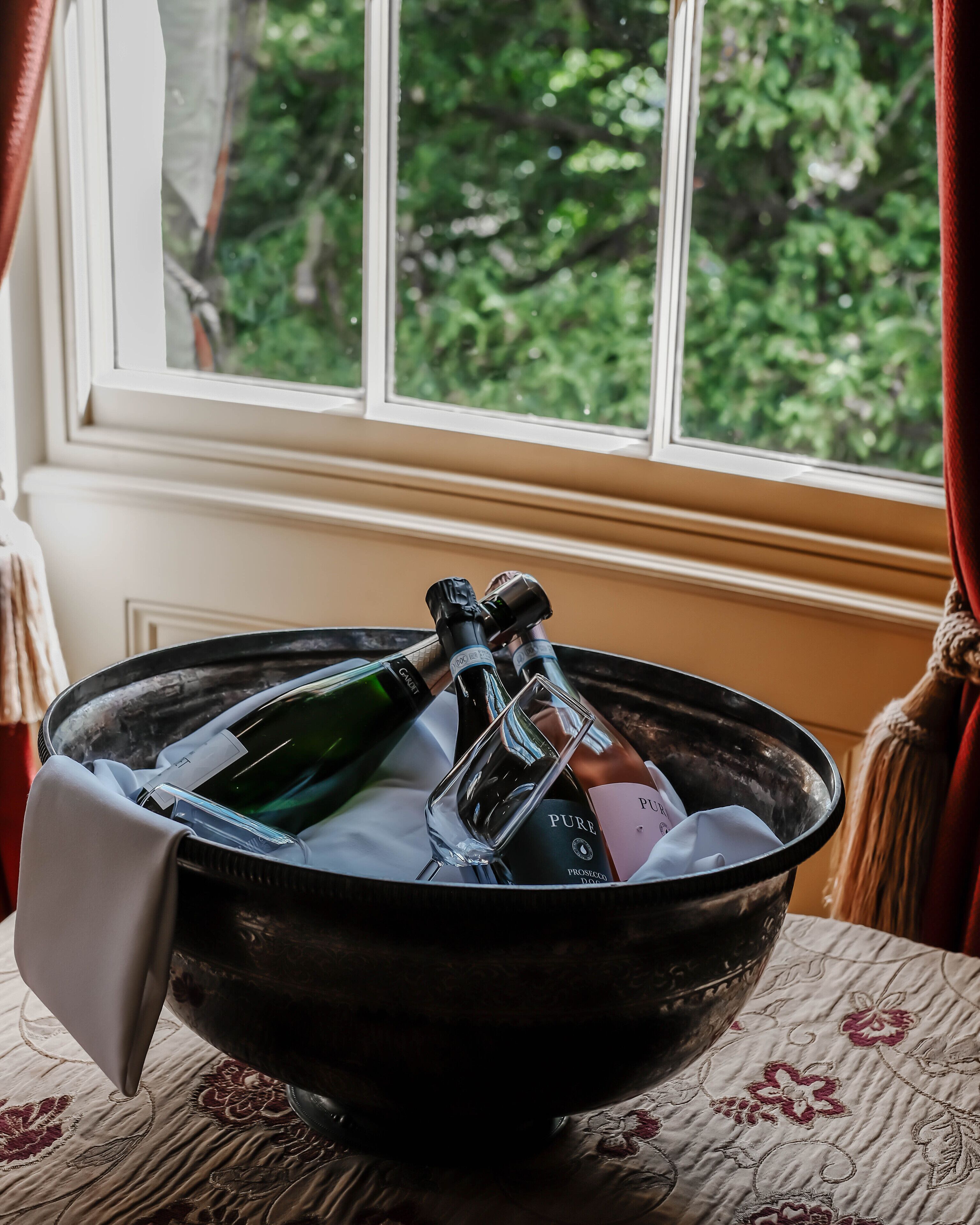 A bowl containing bottles and glasses on a table by a window with a view of trees.