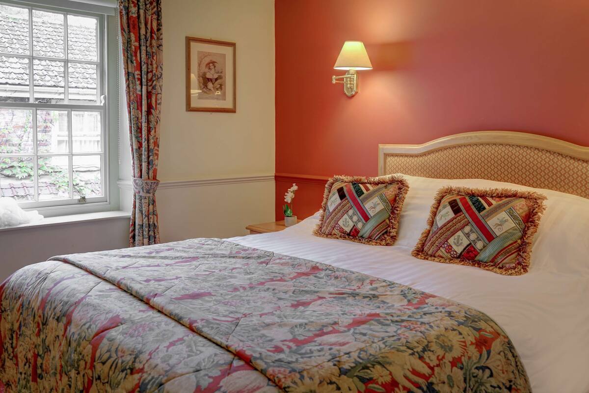 Une chambre à coucher avec une couvre-lit à motifs floraux, une table de chevet avec une lampe et une fenêtre avec des rideaux.