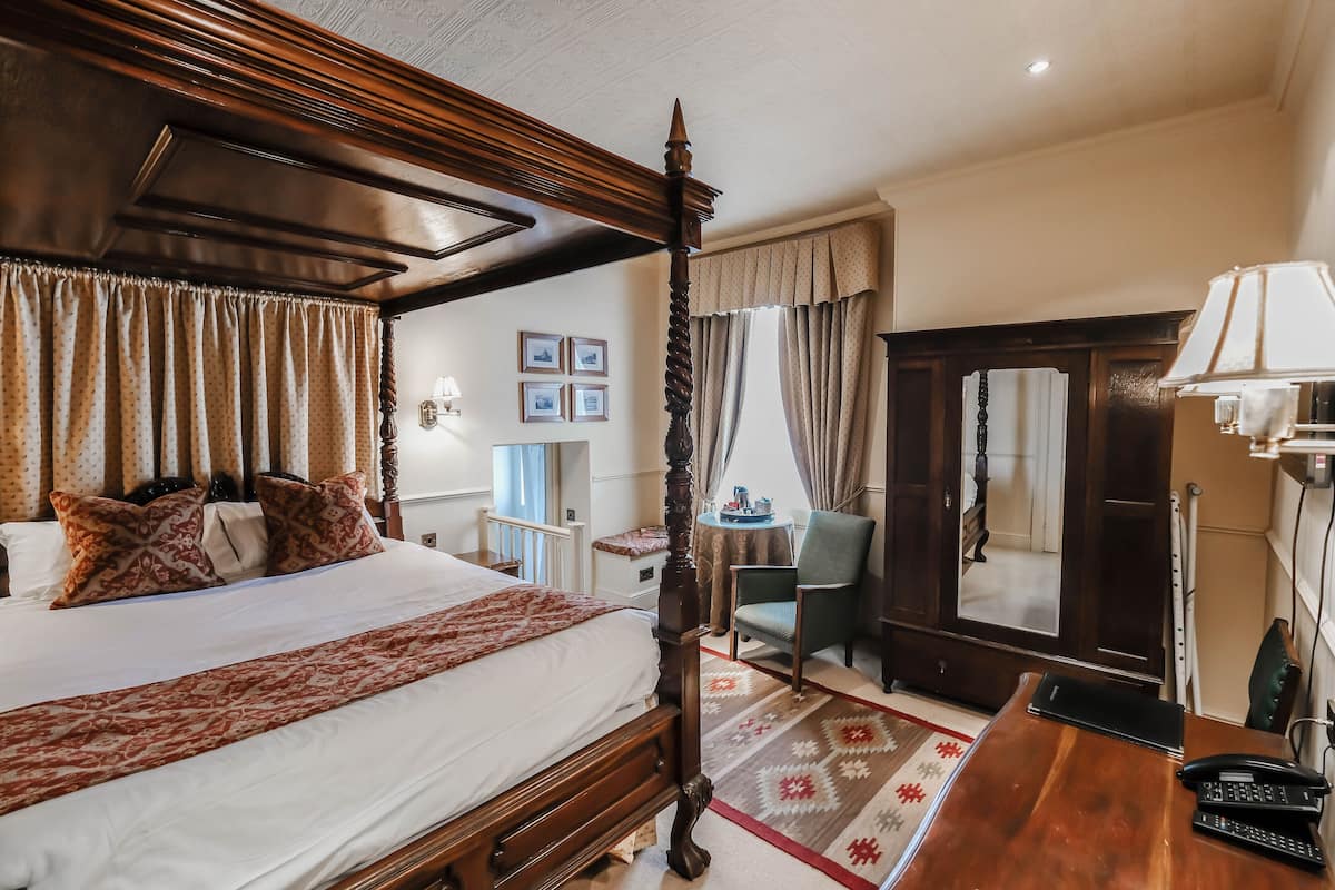 Un lit à baldaquin avec une tête de lit en bois, un bureau avec un téléphone et une chaise, dans une chambre d’hôtel.