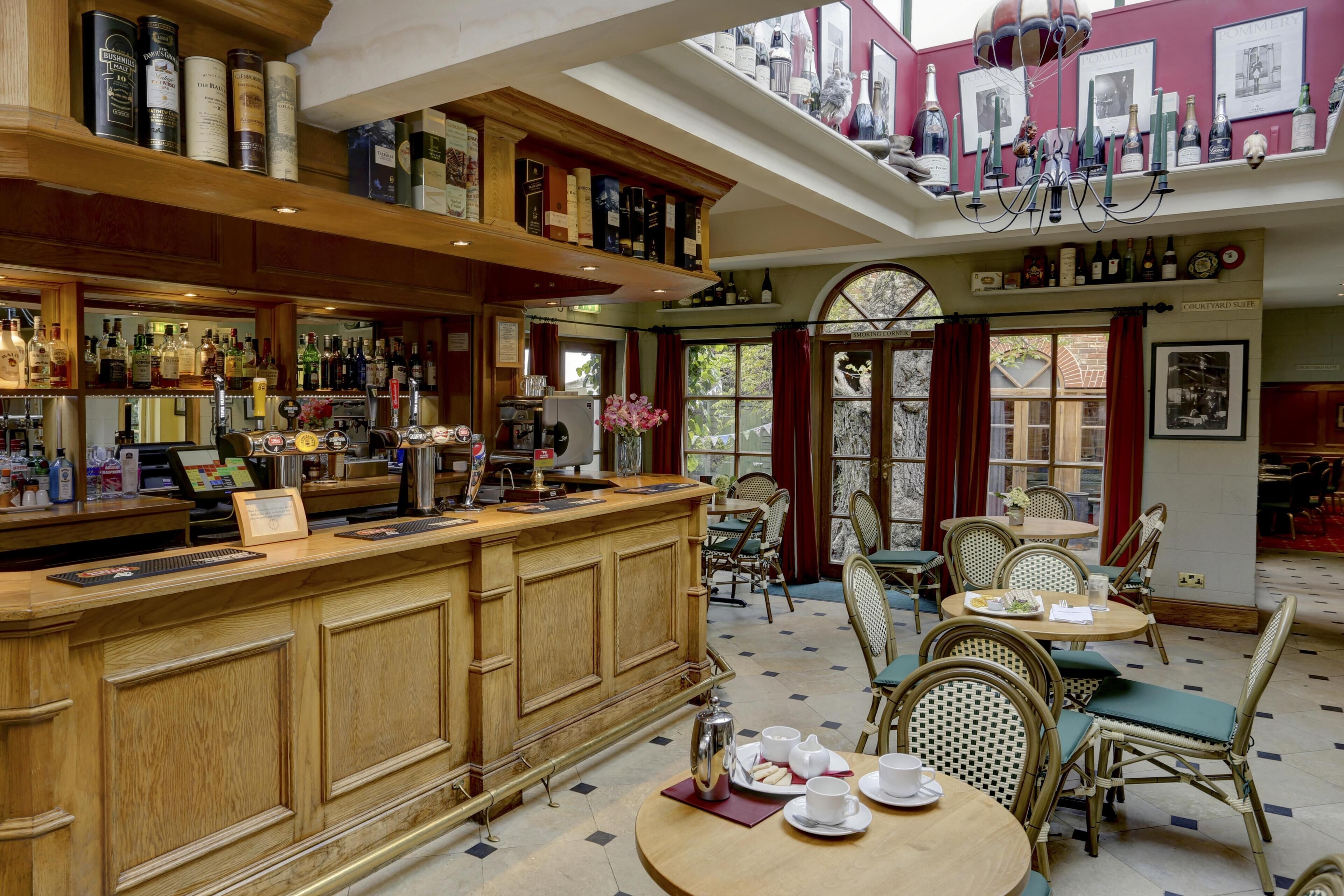 Zona de bar con mostrador y estanterías de madera, mesas de comedor con sillas y vistas a la calle a través de grandes ventanales.