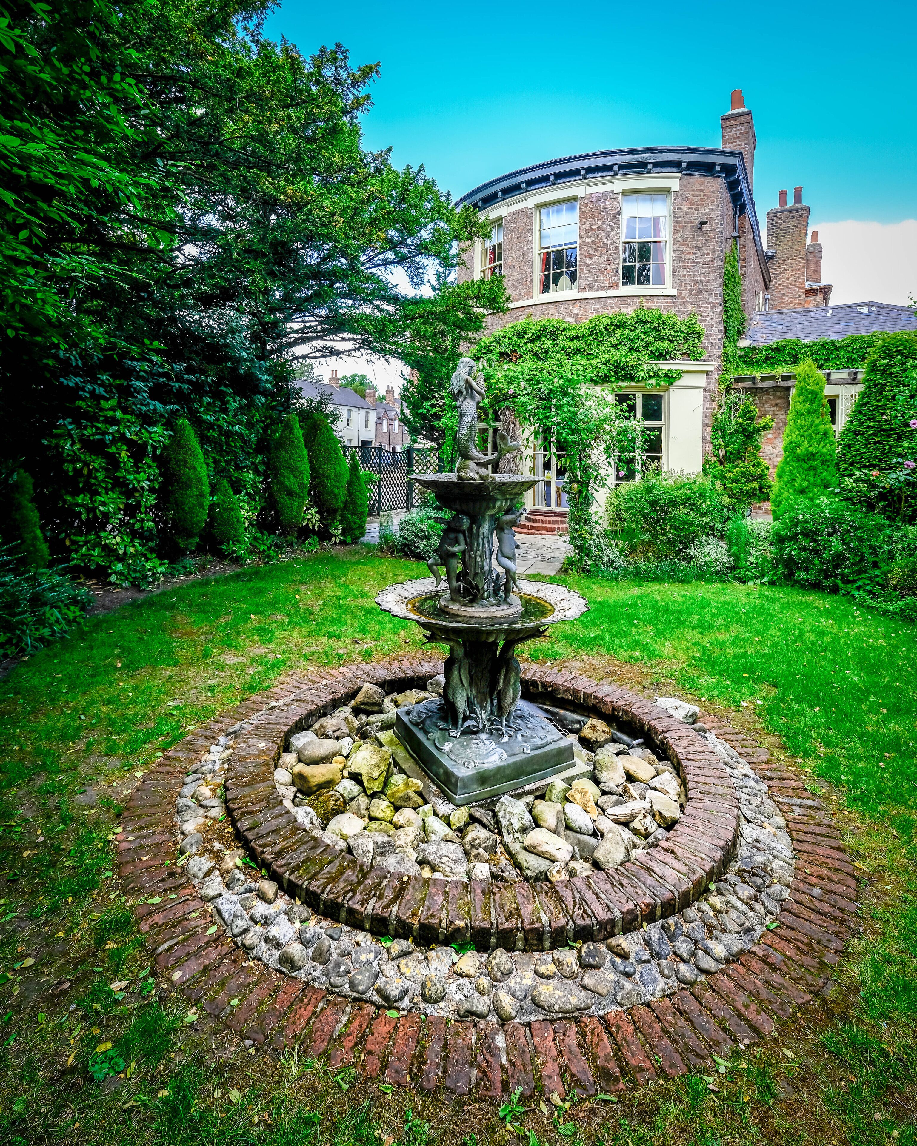A circular brick fountain with a statue in the center, surrounded by a well-maintained garden with hedges and trees.