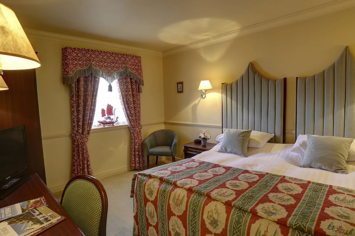 Une chambre d’hôtel comprenant un lit, un bureau avec un ordinateur, une chaise, une fenêtre avec des rideaux et une lampe fixée au mur.