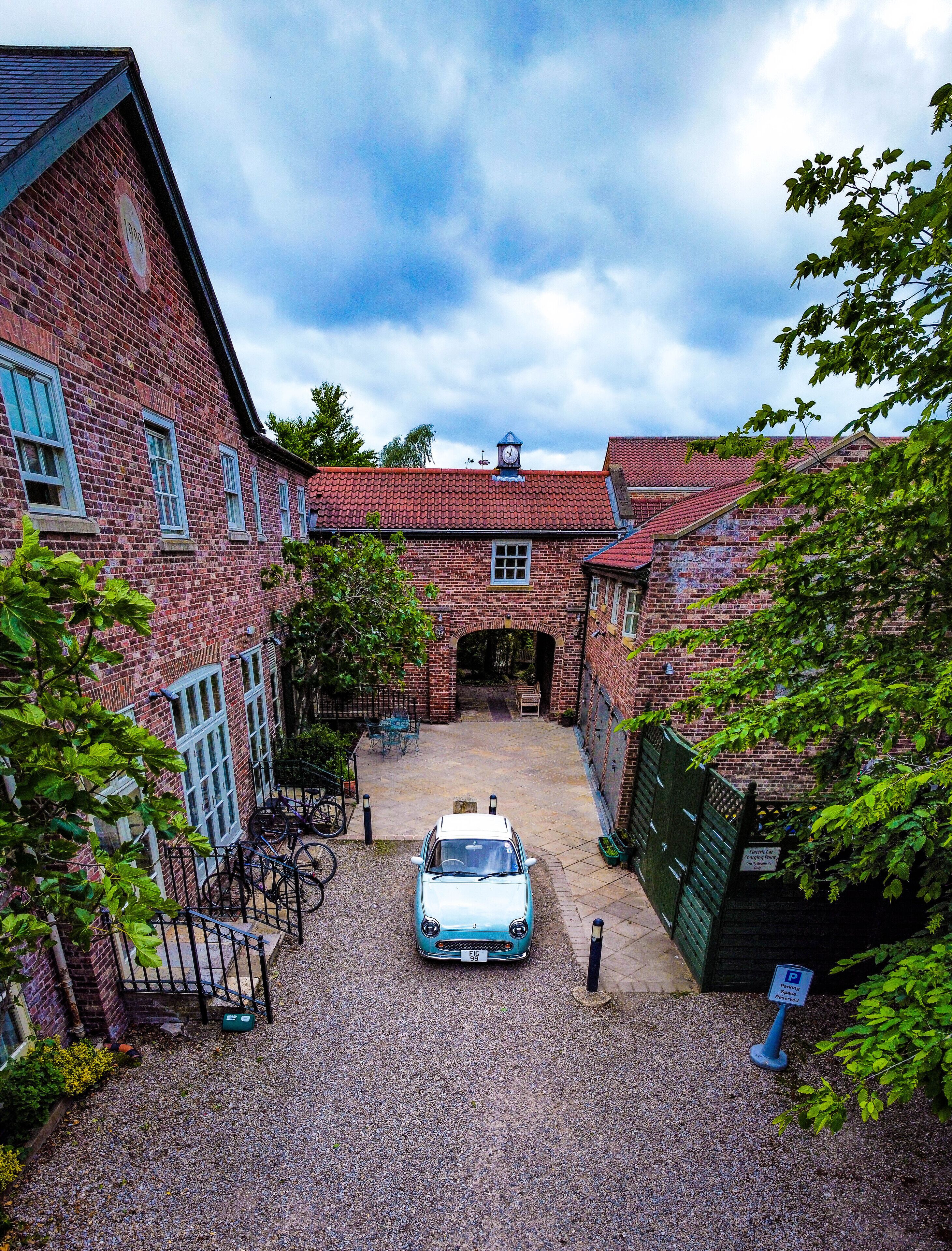 A courtyard with a blue car, brick buildings, and a clock tower.