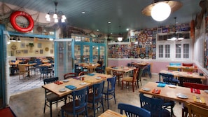 Un restaurante con varias mesas y sillas, decoración náutica con redes y un timón en la pared, y cuadros enmarcados adornando las paredes.