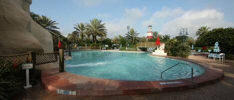 Una piscina con una torre de salvavidas, palmeras y un faro rojo y blanco al fondo.