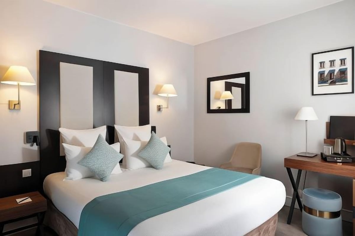Ein Hotelzimmer mit einem großen Bett, zwei Wandlampen, einem Schreibtisch mit Stuhl und einem gerahmten Bild an der Wand.